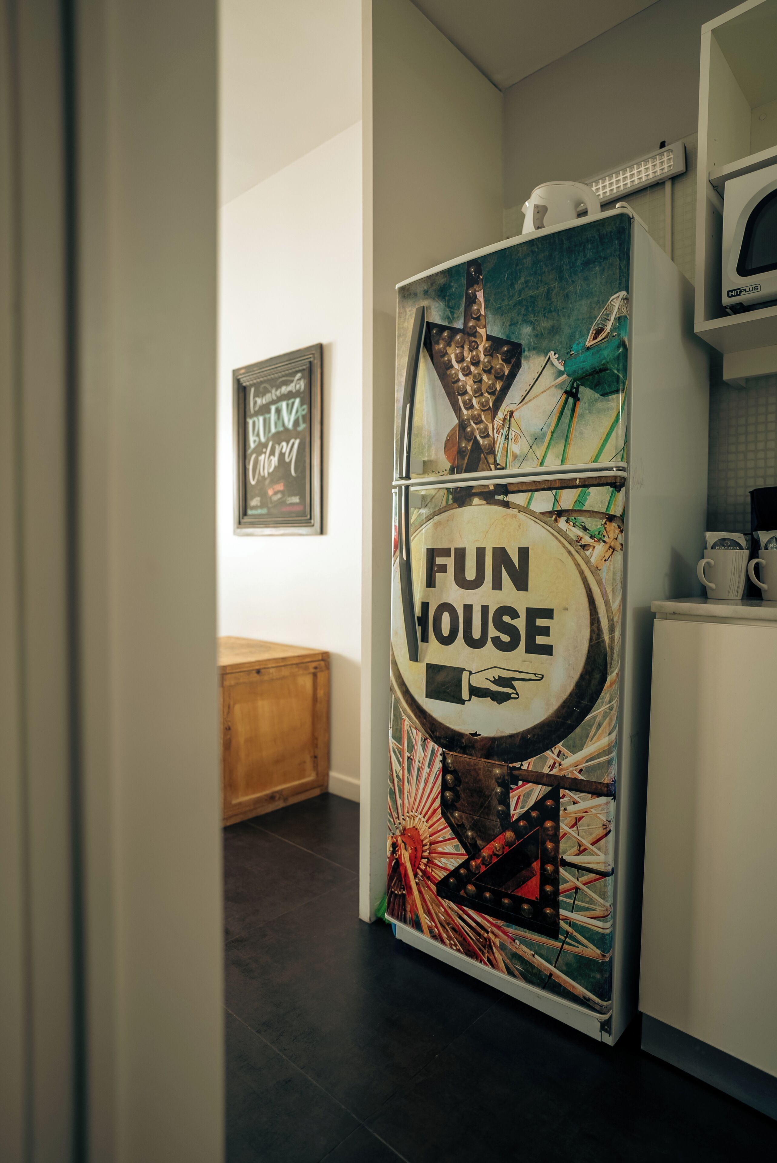 Una cocina con un refrigerador decorado con un letrero que dice 'Fun House', un armario de madera y un cartel en la pared.