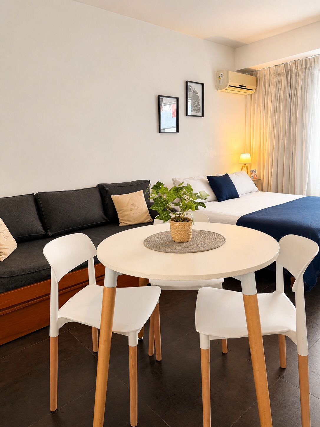 A modern hotel room with a round dining table, white chairs, a black sofa, a bed with a blue blanket, a potted plant, and a wall-mounted air conditioner.