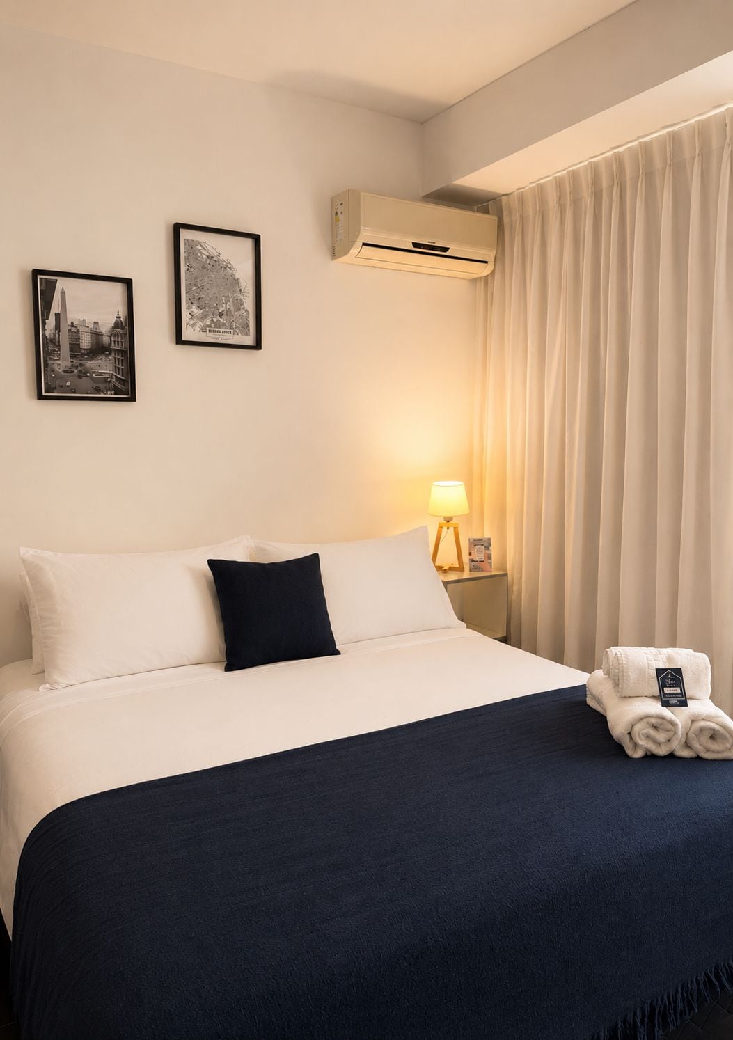 A hotel room with a bed, two framed pictures, an air conditioning unit, a lamp, and a window with curtains.
