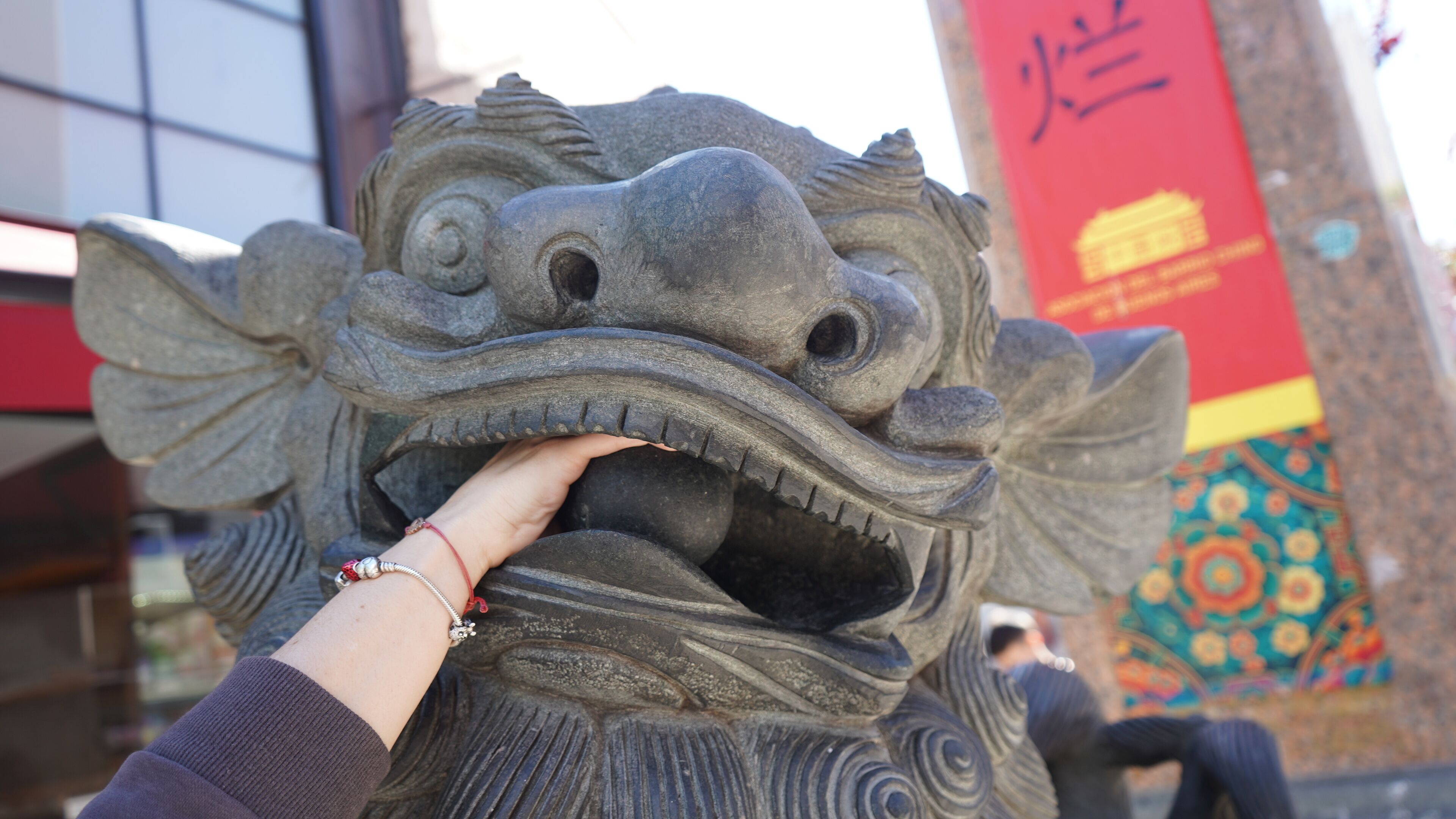 A stone lion statue with intricate carvings, a person touching its mouth, and a colorful mural in the background.