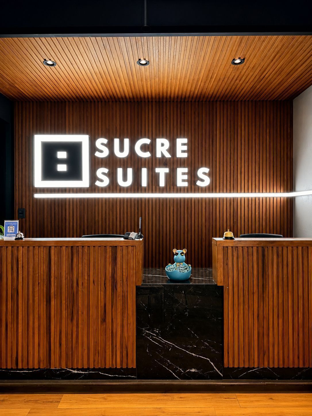 Balcão de recepção com painel de madeira, letreiro de neon com a inscrição 'SUCRE SUITES' e bancada de mármore.