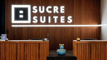 Balcão de recepção com painel de madeira, letreiro de neon com a inscrição 'SUCRE SUITES' e bancada de mármore.