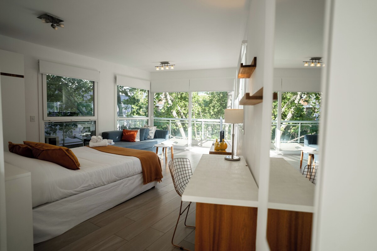 A modern hotel room with a bed, a desk, a chair, and large windows offering a view of the outdoors.