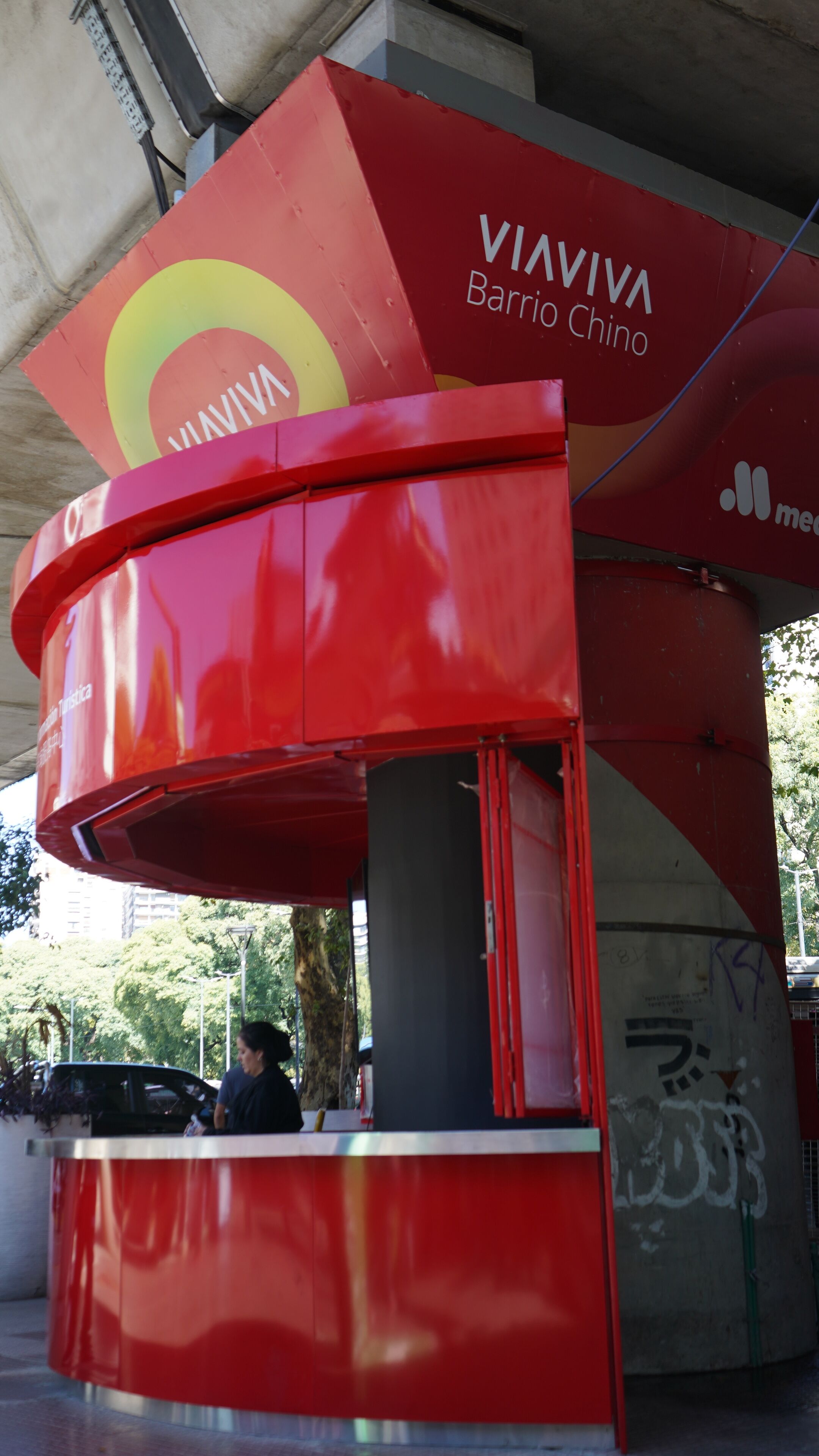 En röd kiosk med texten ”VIAVIVA Barrio Chino” och en del av ett fordon syns i bakgrunden.