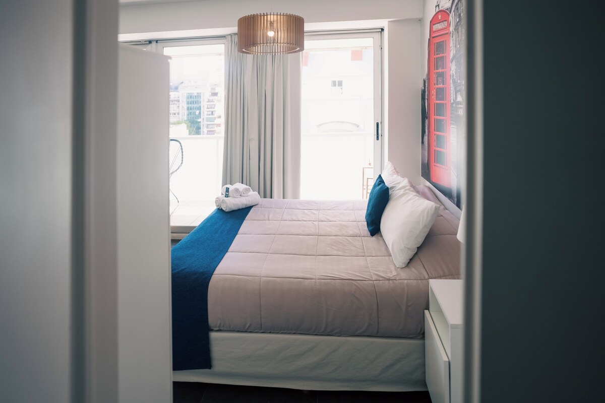 A hotel room with a bed, a window with curtains, a wall-mounted telephone, and a towel on the bed.