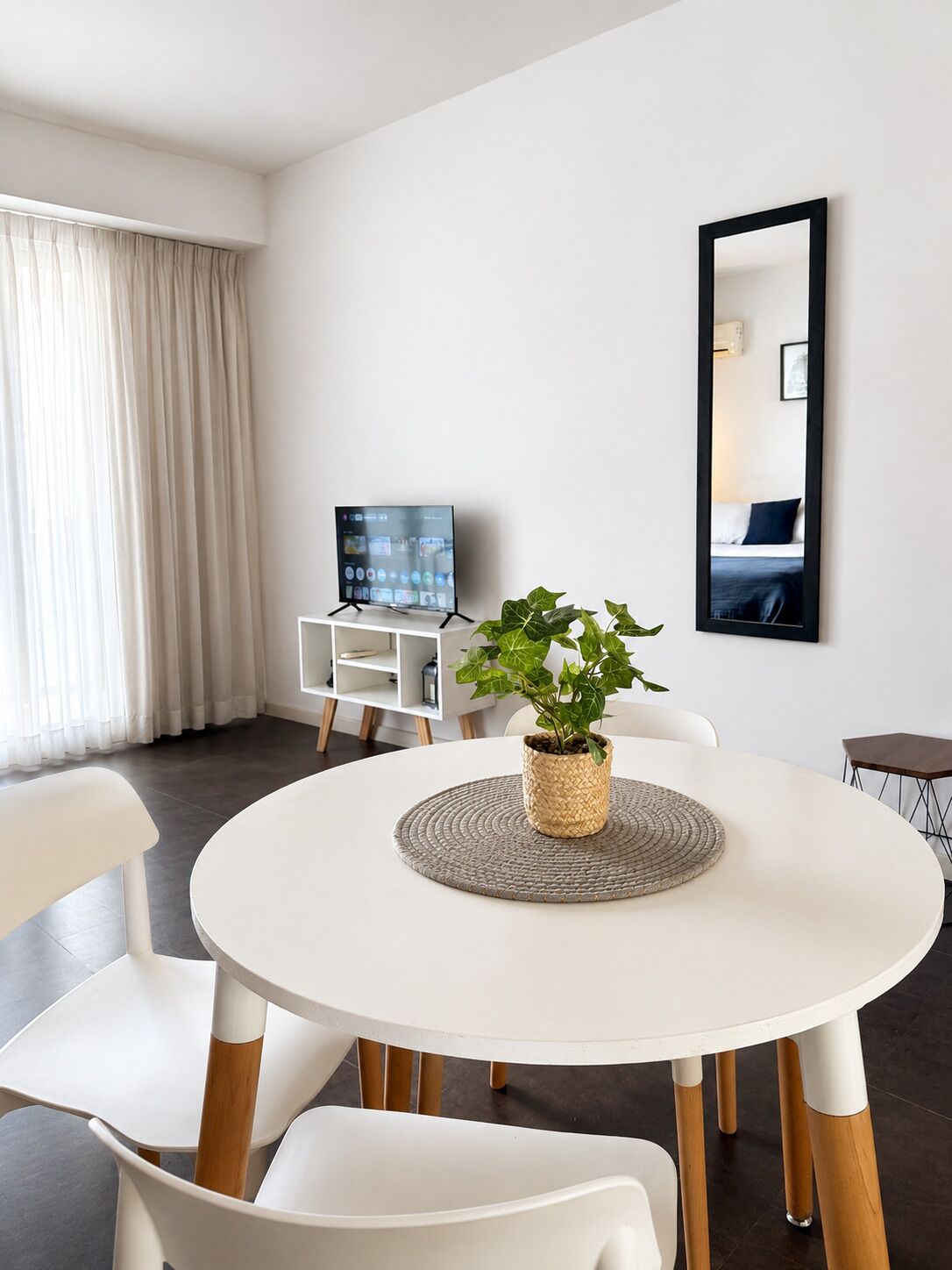 A modern living room with a round dining table, white chairs, a potted plant, a flat-screen TV, and a large mirror on the wall.