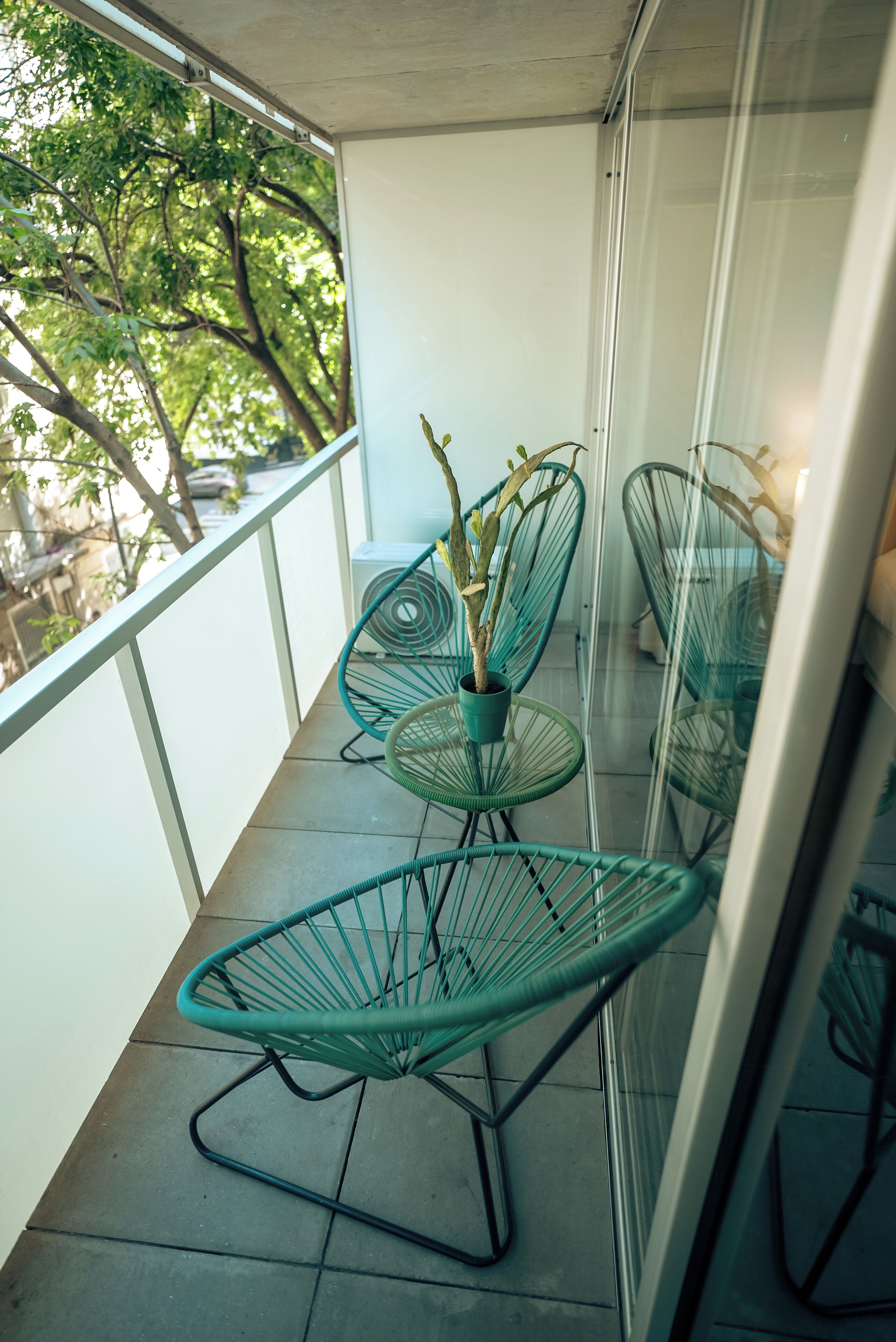 Un balcón con barandilla de vidrio, una planta en maceta y dos sillas de estructura metálica verde.