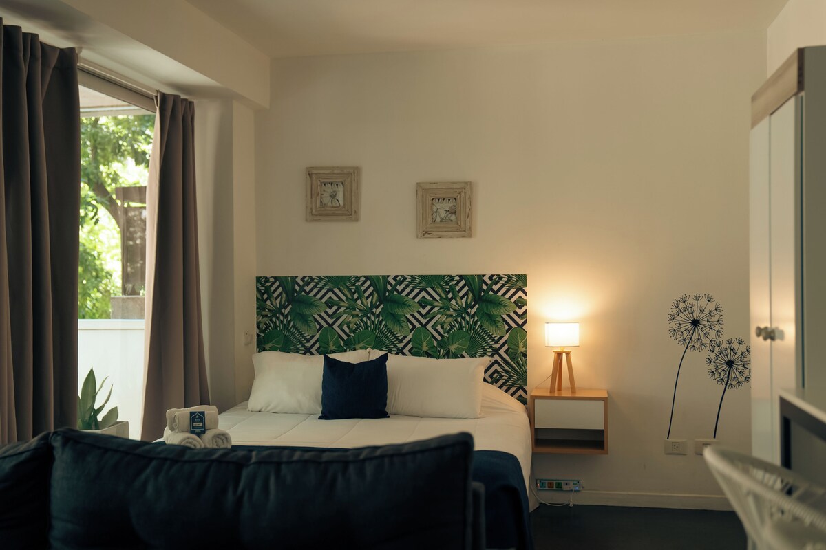 A hotel room with a bed, a bedside table with a lamp, a wall with framed pictures, and a window with curtains.