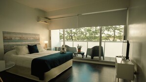 A modern hotel room with a large bed, a small table with a chair, a potted plant, and a view of trees through large windows. - Sucre Suites (Buenos Aires)