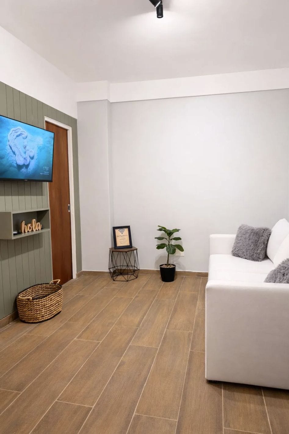 A living room with a white sofa, a flat-screen TV mounted on a wall, a potted plant, a framed certificate, and a woven basket.