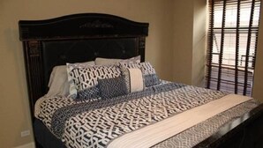 Uma cama com colcha estampada e almofadas, uma cabeceira de madeira escura e uma janela com persianas.