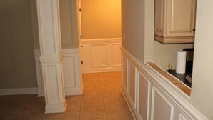 Um corredor com piso ladrilhado, rodapé branco e uma porta que dá para um quarto com parede bege e luminária.