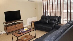 Uma sala de estar com um sofá de couro preto, uma mesa de centro de madeira, uma TV sobre um suporte de madeira e uma janela com persianas.