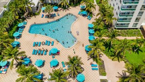 Vista aérea de uma área de piscina de hotel, com várias espreguiçadeiras e guarda-sóis, cercada por palmeiras e uma piscina.