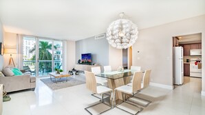Uma sala de estar moderna com uma mesa de jantar de vidro, cadeiras brancas, um sofá e uma grande janela com vista para palmeiras e edifícios.