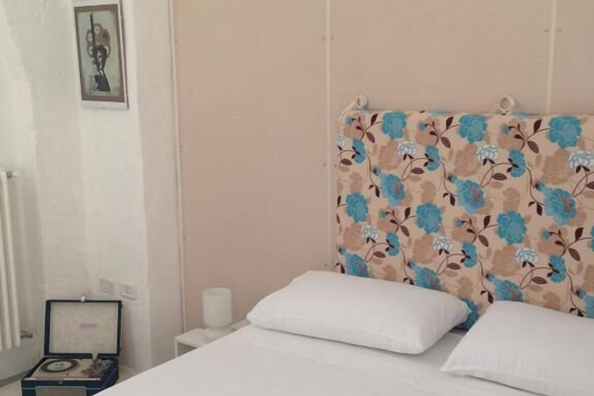 Ein Bett mit weißer Bettwäsche, ein Kopfseitenverkleidung mit Blumenmuster und ein kleiner Nachttisch mit einem Radio.