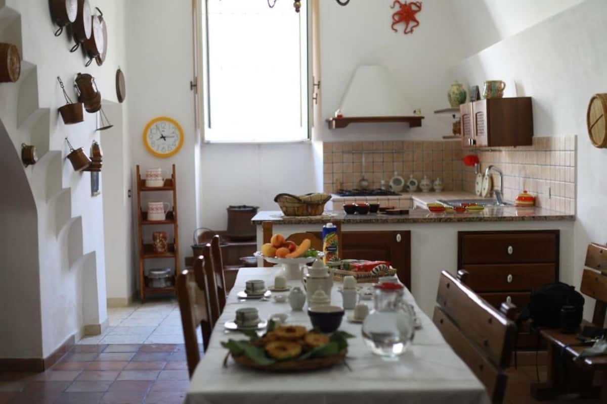 Eine Küche mit Esstisch, gedeckt für eine Mahlzeit, einer Wanduhr sowie verschiedenen Küchenutensilien und Geräten.