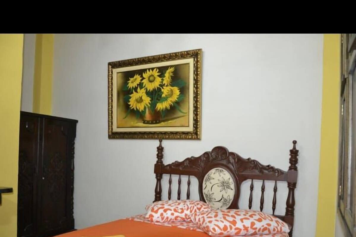 A bedroom with a wooden bed, a floral pillow, and a framed painting of sunflowers on the wall.