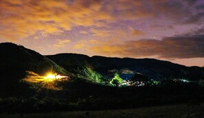 Escena nocturna con cielo estrellado, una colina con una estructura pequeña iluminada y un árbol en primer plano.