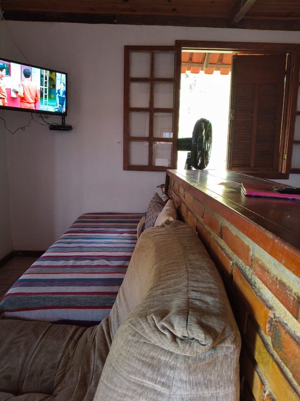 A room with a bed, a television mounted on the wall, a window with wooden shutters, and a fan on the window sill.
