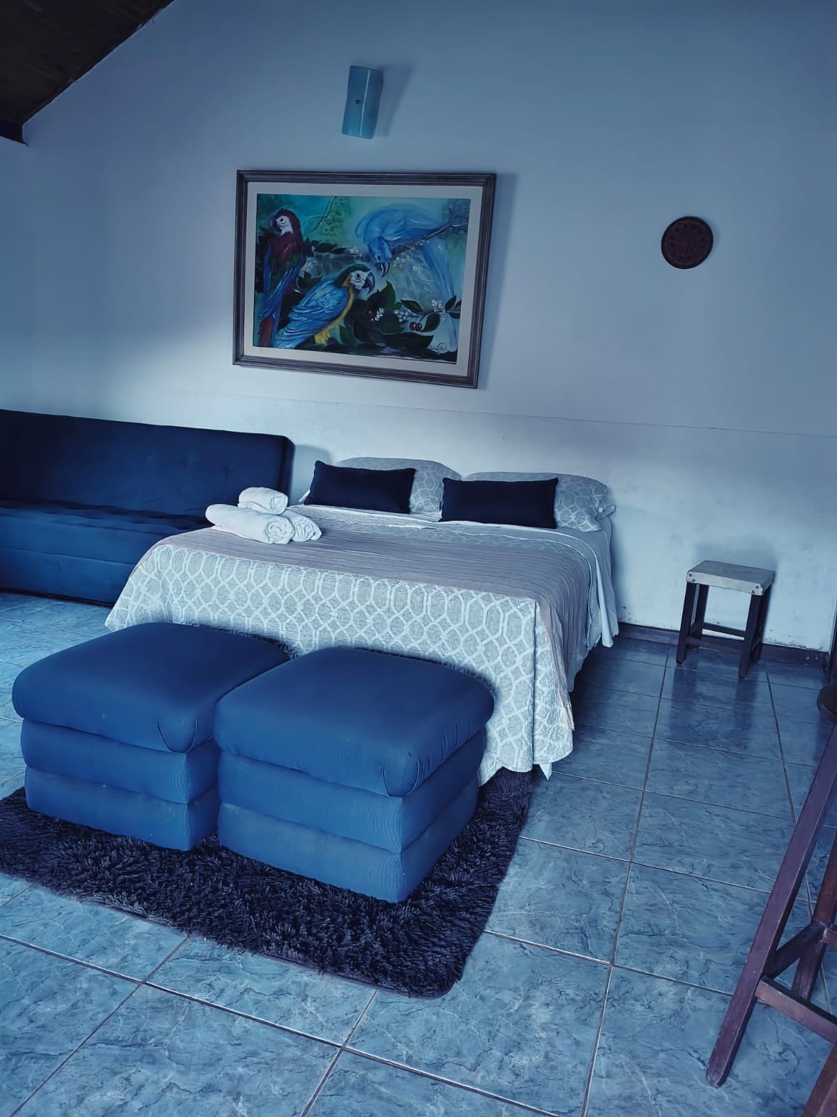 A bedroom with a bed, a sofa, a small stool, and a painting on the wall.