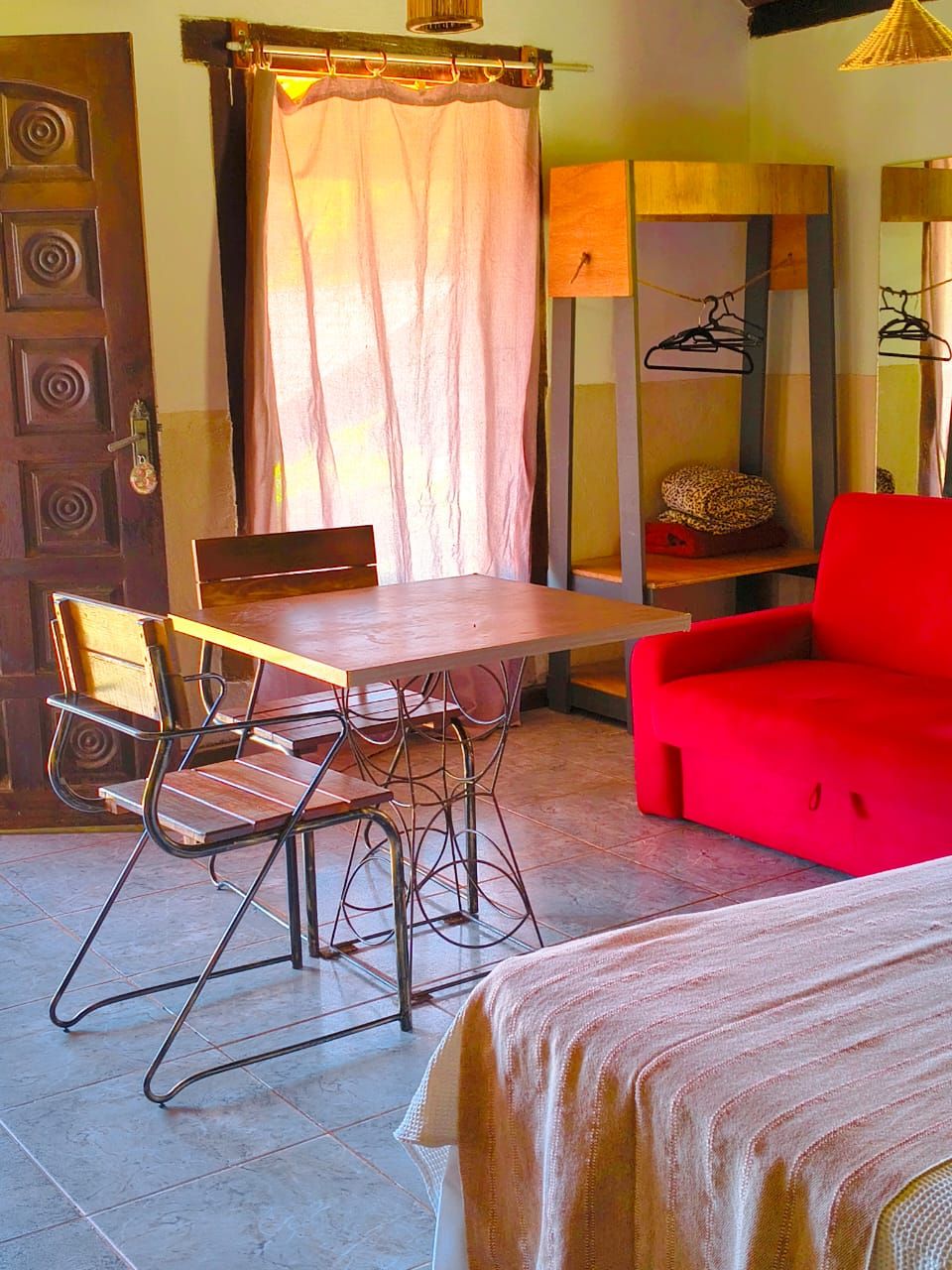 A room with a wooden table, metal chairs, a red sofa, a bed, a wooden cabinet, and a hanging clothes rack.