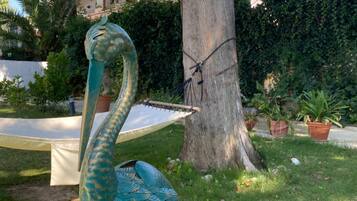 A sculpture of a green bird with a long neck and tail, positioned in a grassy area with a tree and potted plants in the background.