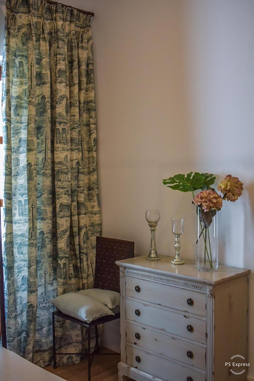 Una habitación con cortina estampada, una silla pequeña, un aparador con cajones y un jarrón con flores.