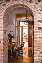 Una habitación con paredes de ladrillo visto, una puerta de madera, un candelabro, un escritorio con un jarrón de flores y una silla.