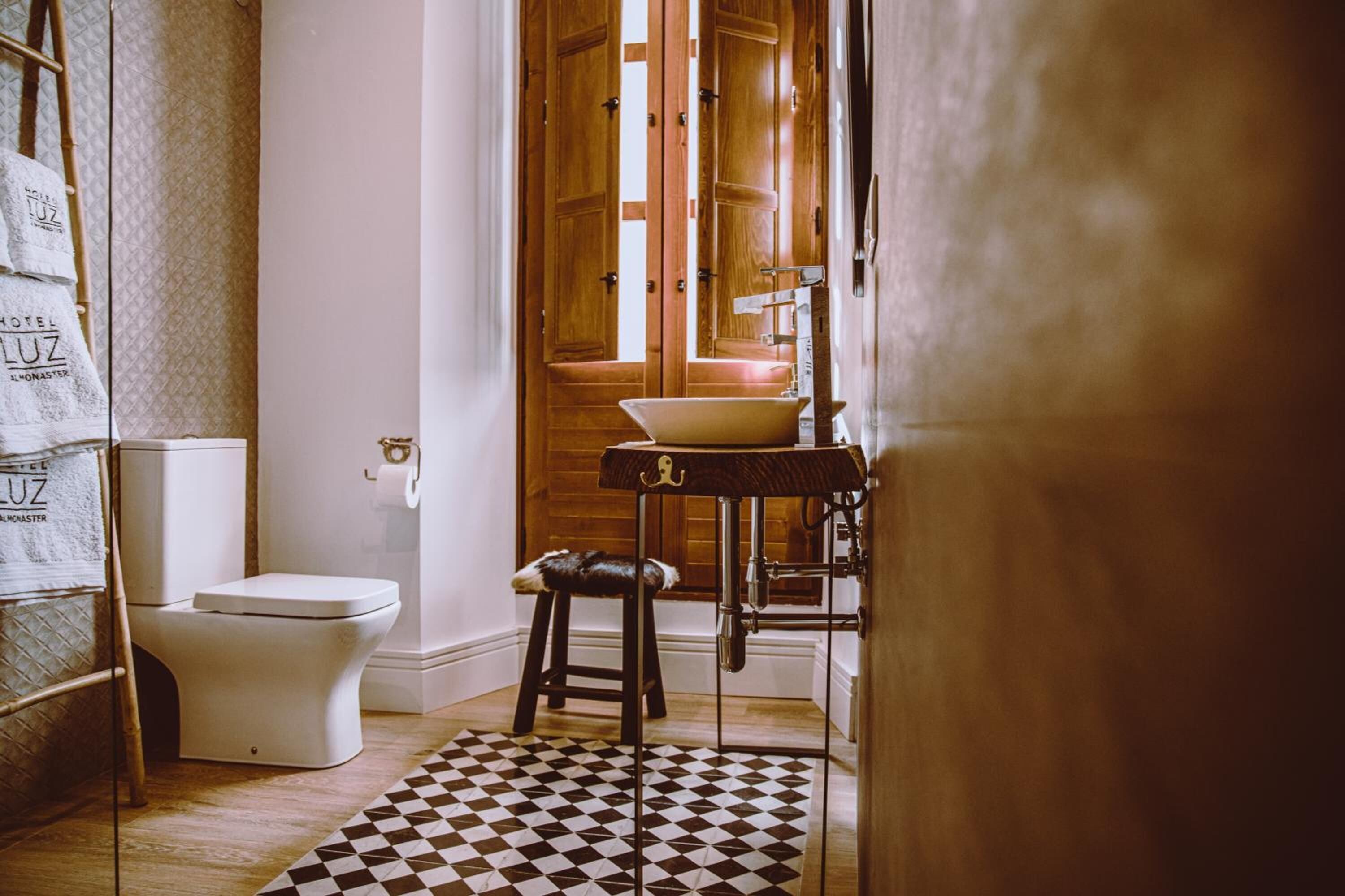 Une salle de bain comprenant des toilettes, un lavabo avec un bac, un tabouret, un sol à carreaux et une porte en bois avec une fenêtre.