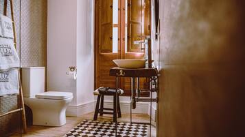 Une salle de bain comprenant des toilettes, un lavabo avec un bac, un tabouret, un sol à carreaux et une porte en bois avec une fenêtre.