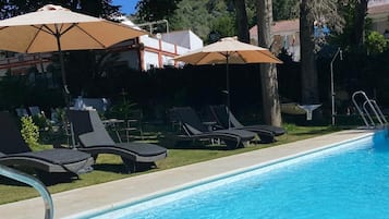 A swimming pool with lounge chairs and umbrellas, surrounded by trees and a building.