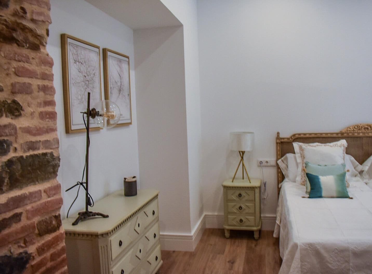 Una camera da letto con parete in mattoni, una cassettiera, un comodino con una lampada, un letto con cuscini e due quadri appesi al muro.