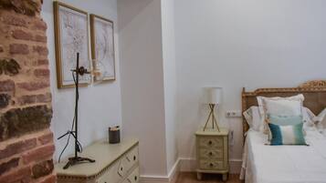 Una camera da letto con parete in mattoni, una cassettiera, un comodino con una lampada, un letto con cuscini e due quadri appesi al muro.