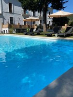 Una piscina con sillas de descanso y sombrillas, rodeada por un edificio con balcones y un árbol.
