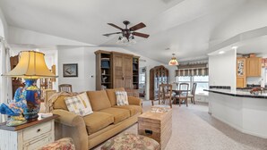 A spacious living room with a sofa, ottoman, and a coffee table. There is a dining area with chairs and a table. A kitchen area is visible with cabinets and a countertop. - BellaVida B&B and Retreat (Wimberley)