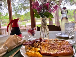 Una mesa de desayuno con un plato de huevos, tostadas y tocino, una jarrón de agua, un florero con flores y una jarra de agua roja.