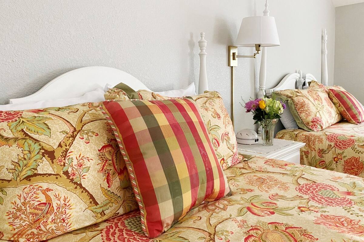 Een keurig opgemaakt bed met bloemenbeddels, een nachtkastje met een lamp en een vaas met bloemen.