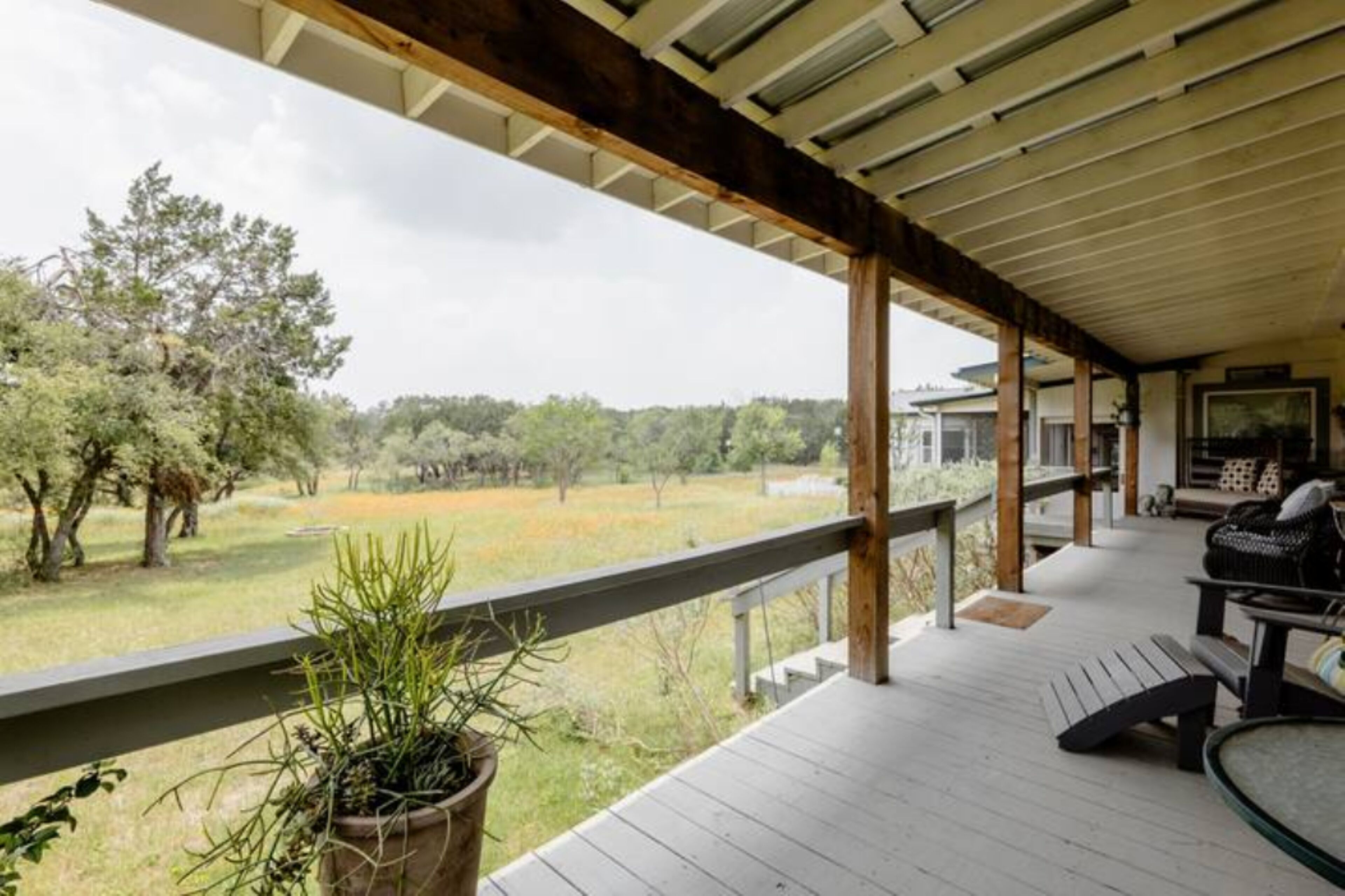 Un porche cubierto con terraza de madera, una planta en maceta, un banco y vistas a un campo cubierto de césped y árboles.