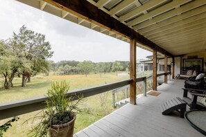 Un porche cubierto con terraza de madera, una planta en maceta, un banco y vistas a un campo cubierto de césped y árboles.