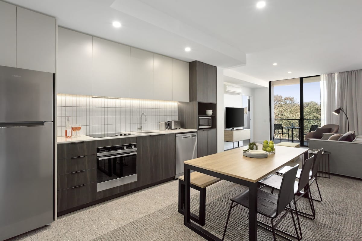 A modern kitchen with grey cabinetry, a wooden dining table, and a view of the outdoors through large windows.