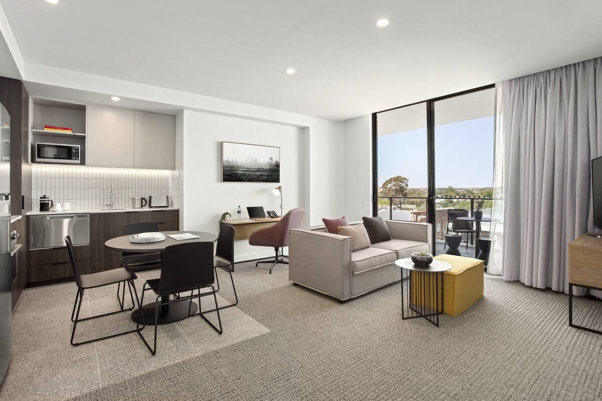 A modern hotel room with a kitchenette, a dining area, a sofa, a TV, and a large window with a view.