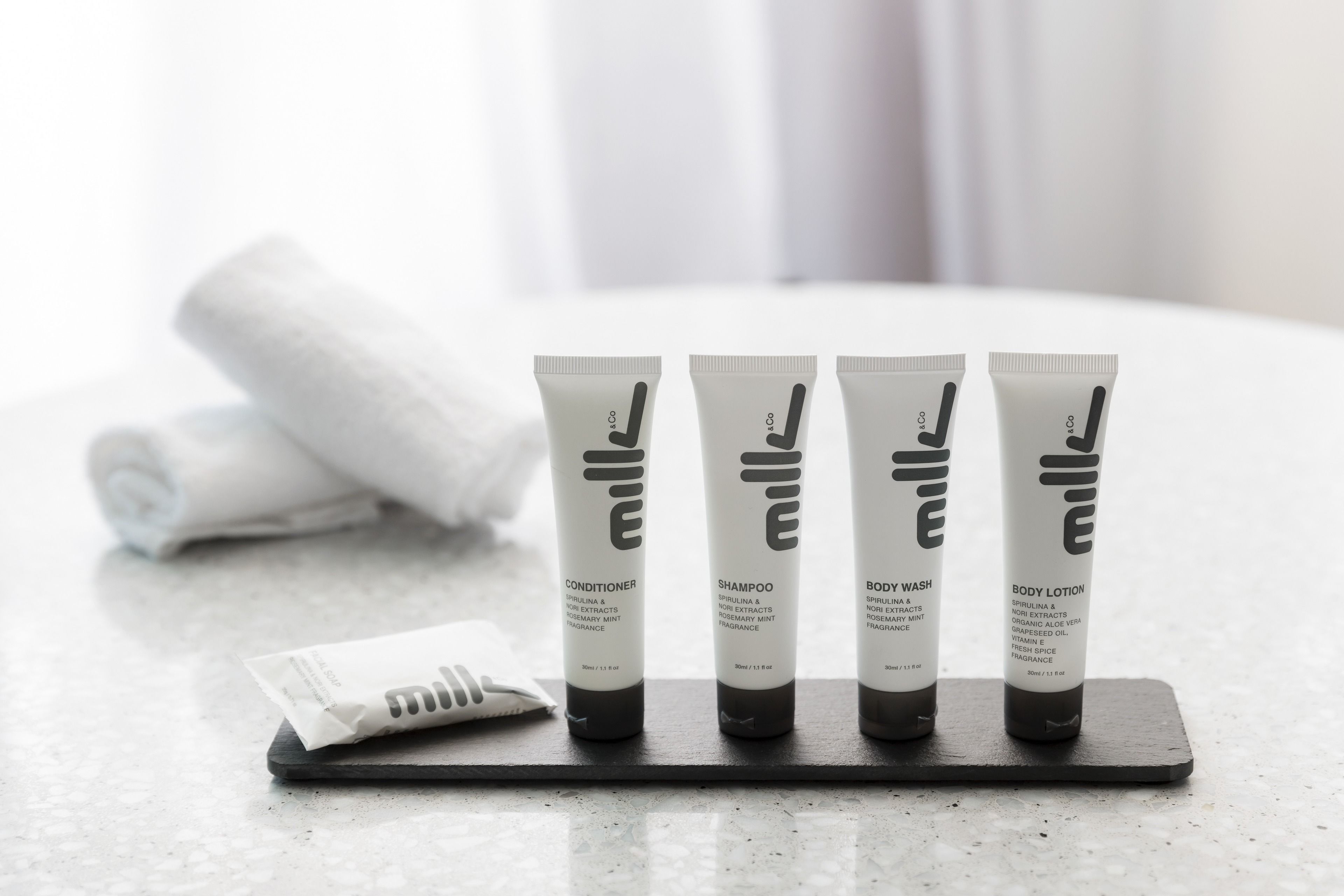 Four toiletry tubes on a tray with a rolled towel in the background.