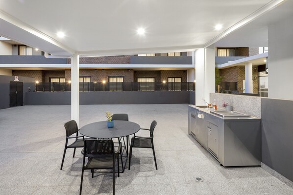 Un patio moderno al aire libre con una mesa redonda y sillas, una zona de cocina integrada y un fondo de pared de ladrillo.