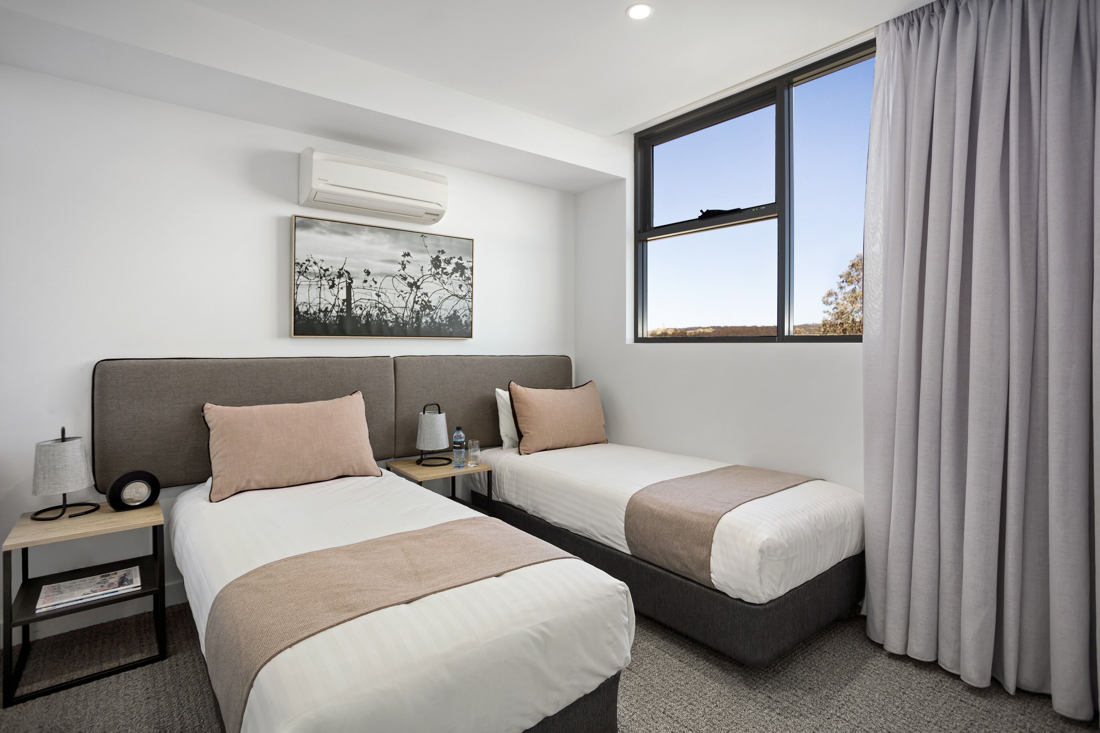 Habitación de hotel con dos camas, una mesita de noche con lámpara, un cuadro en la pared y una ventana con cortinas.
