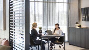 A modern office space with two people seated at a table with laptops, a large window with blinds, and a mounted flat-screen TV on the wall.