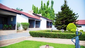A building with a red-tiled roof, a covered outdoor seating area, and a well-maintained garden with hedges and a statue. - Rexmar Hotel (Kumasi)