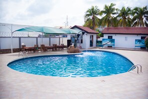 A swimming pool with a stone border, a covered seating area with chairs, and a small building with a red roof. - Rexmar Hotel (Kumasi)