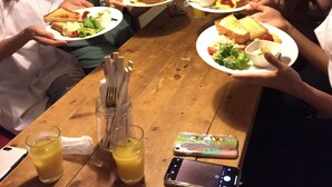 A group of people sitting around a wooden table with plates of food, a glass of juice, and a smartphone on the table. - Heart Hostel and Diner (Ise)