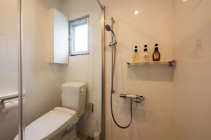 A bathroom with a toilet, a shower with a handheld showerhead, and a shelf with three bottles. - Tsukasa Condo Chatan Araha (Chatan)